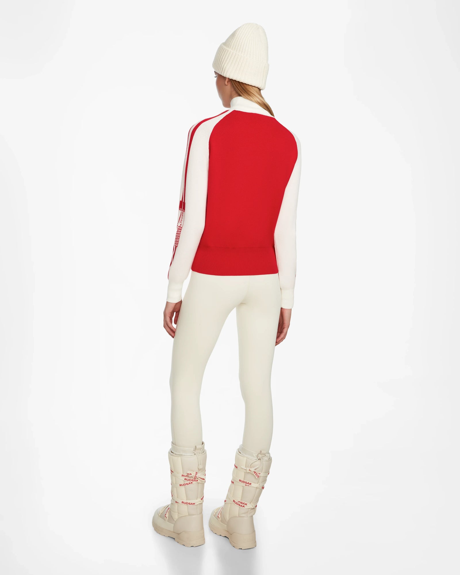 A person stands facing away in the Rudsak FANNY - 8524506 Flame Red après-ski sweater, white leggings, a white beanie, and chunky white winter boots with red accents against a plain white background.
