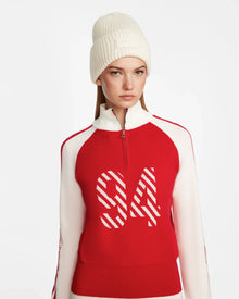A woman in a white knit beanie and the Rudsak FANNY - 8524506 Flame Red après-ski sweater with bold 94 stripes and a mock-neck zip-up stands against a plain white background.