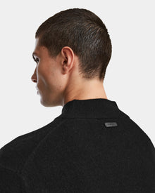 A person with short brown hair is shown from the back and side, modeling the Rudsak FERRAN - 6524511 Black sweater against a plain white background. A small rectangular black tag appears on the upper back of the sweater.