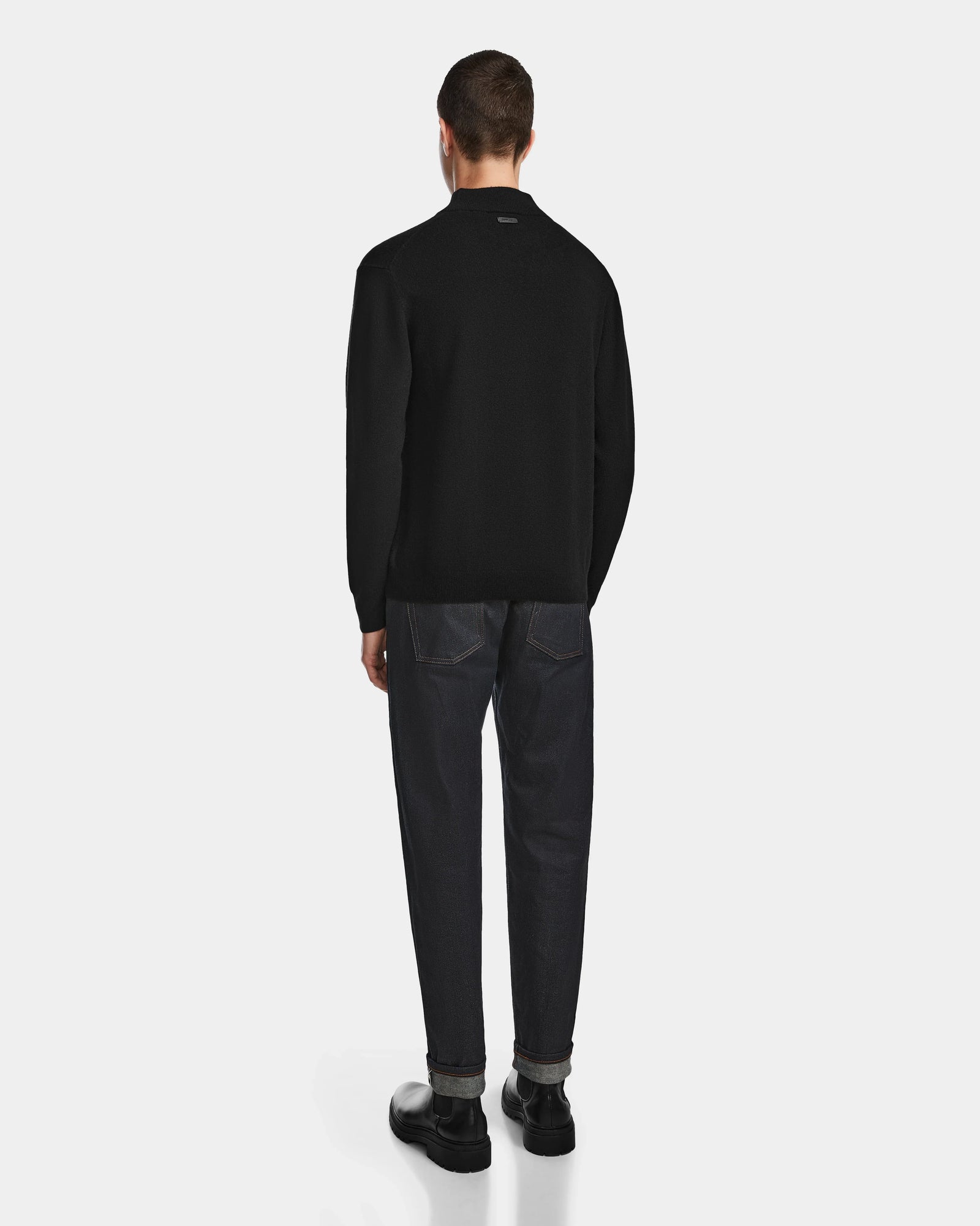 A person stands facing away from the camera, wearing the Rudsak FERRAN - 6524511 Black mens turtleneck sweater, paired with dark jeans and black shoes against a plain white background.