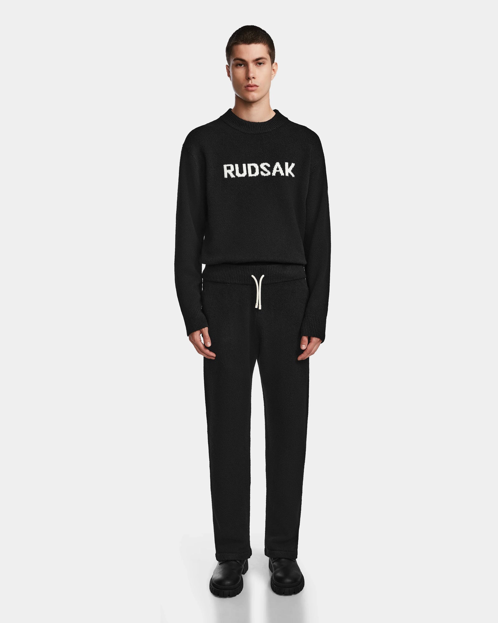 A young man faces forward wearing the Rudsak GEIGER - 9524512 Black M sweater with white lettering, matching relaxed fit lounge pants with a white drawstring, and black shoes, all set against a plain white background.