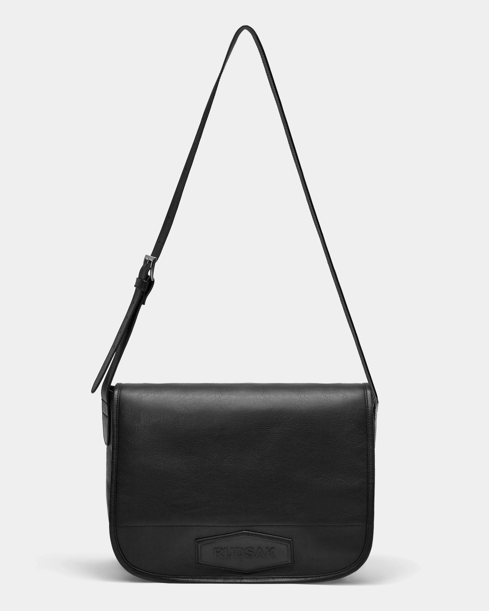 The Rudsak GRAYSON L - 8324525 Black is a black leather crossbody bag with a rectangular flap, long adjustable strap, and subtle embossed logo on the front. Simple and elegant, its shown against a plain white background.