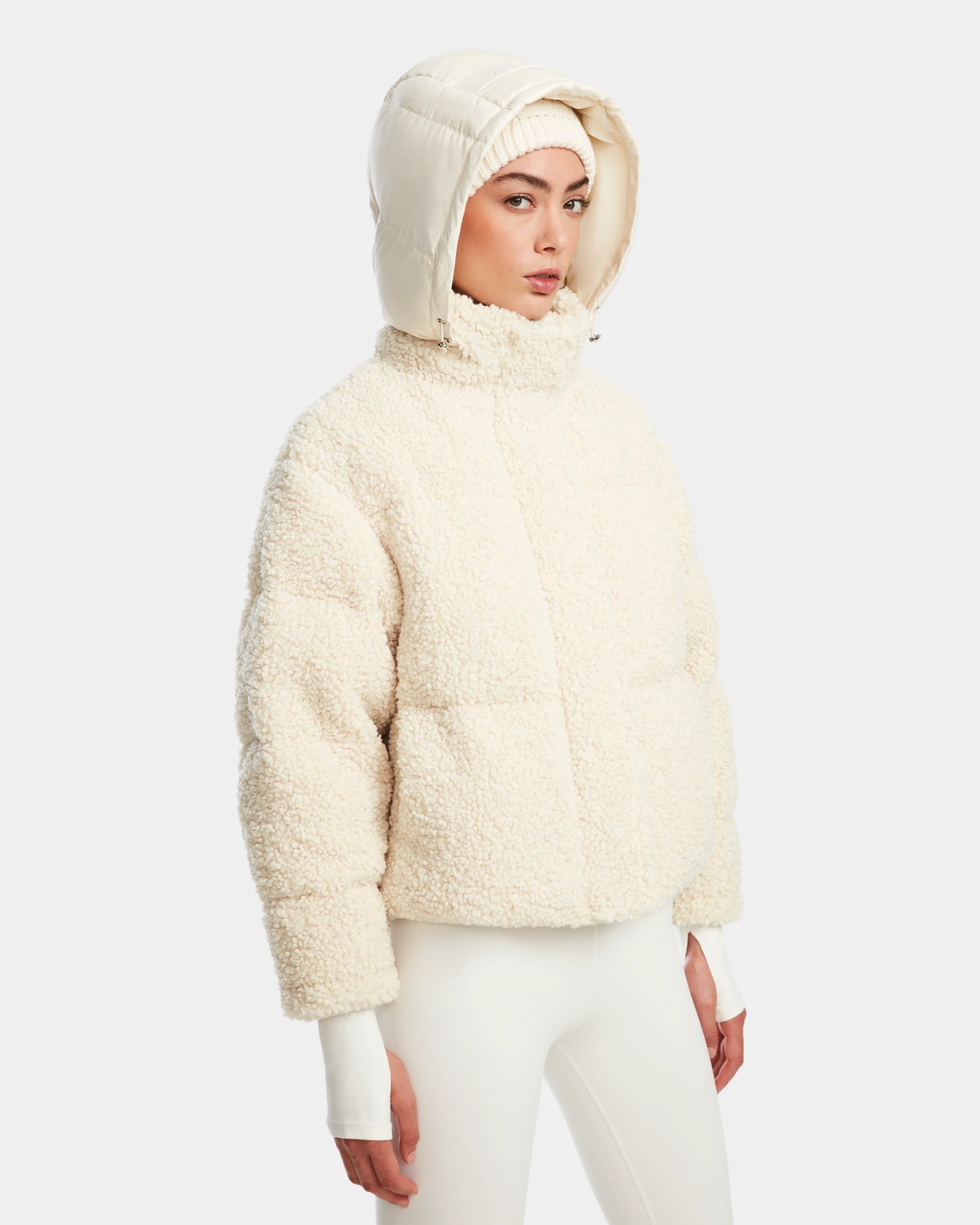 A woman in the Rudsak IKSA - 8124706 Iceland off-white teddy down puffer jacket with a matching hood and white pants stands against a plain background, looking slightly to the side with a neutral expression.