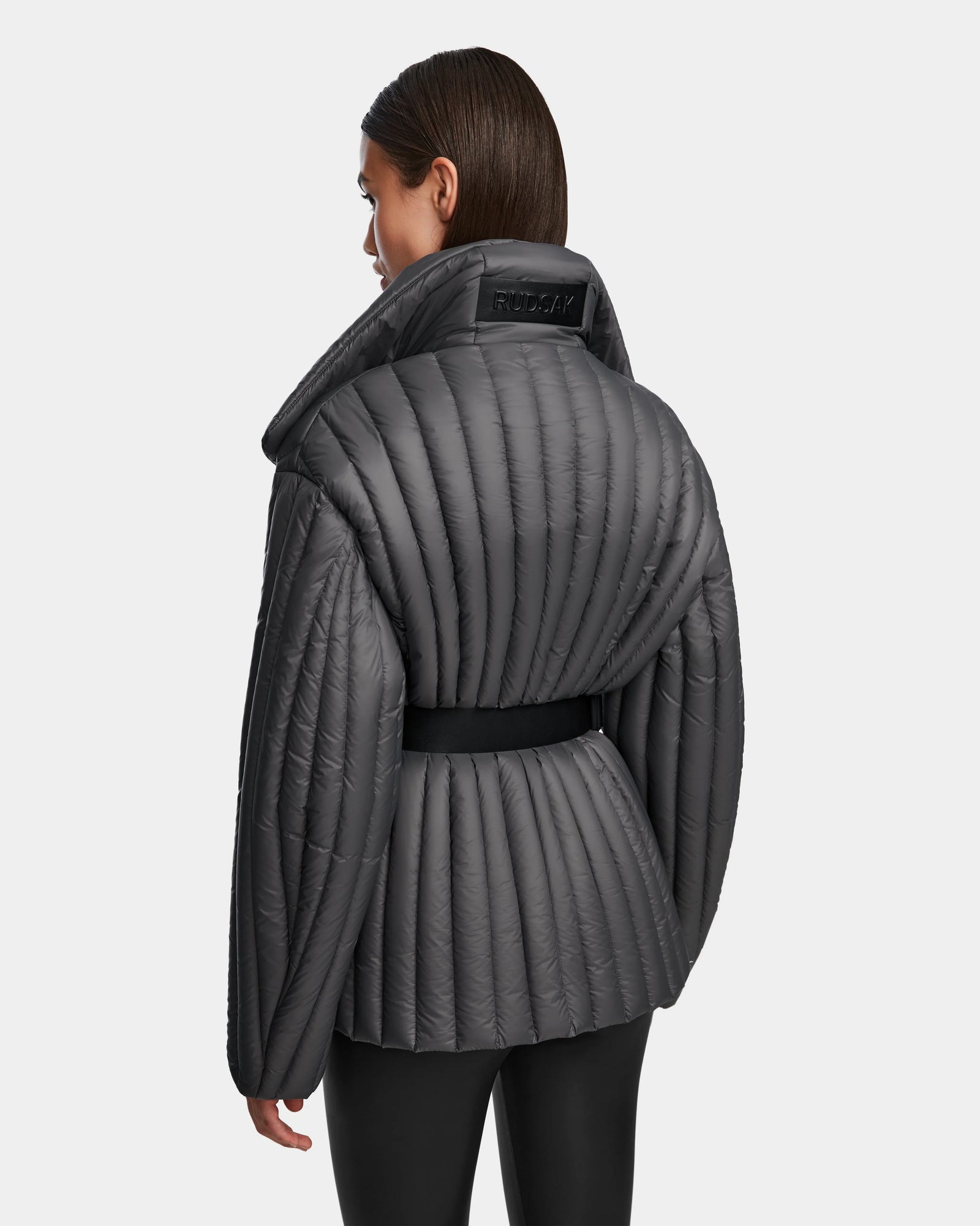 A woman with straight brown hair models the Rudsak INES T - 8124625 Grey, a dark gray womens light down jacket with heavy quilting, a high wide collar, and a belted waist. She faces away to show its stylish back.