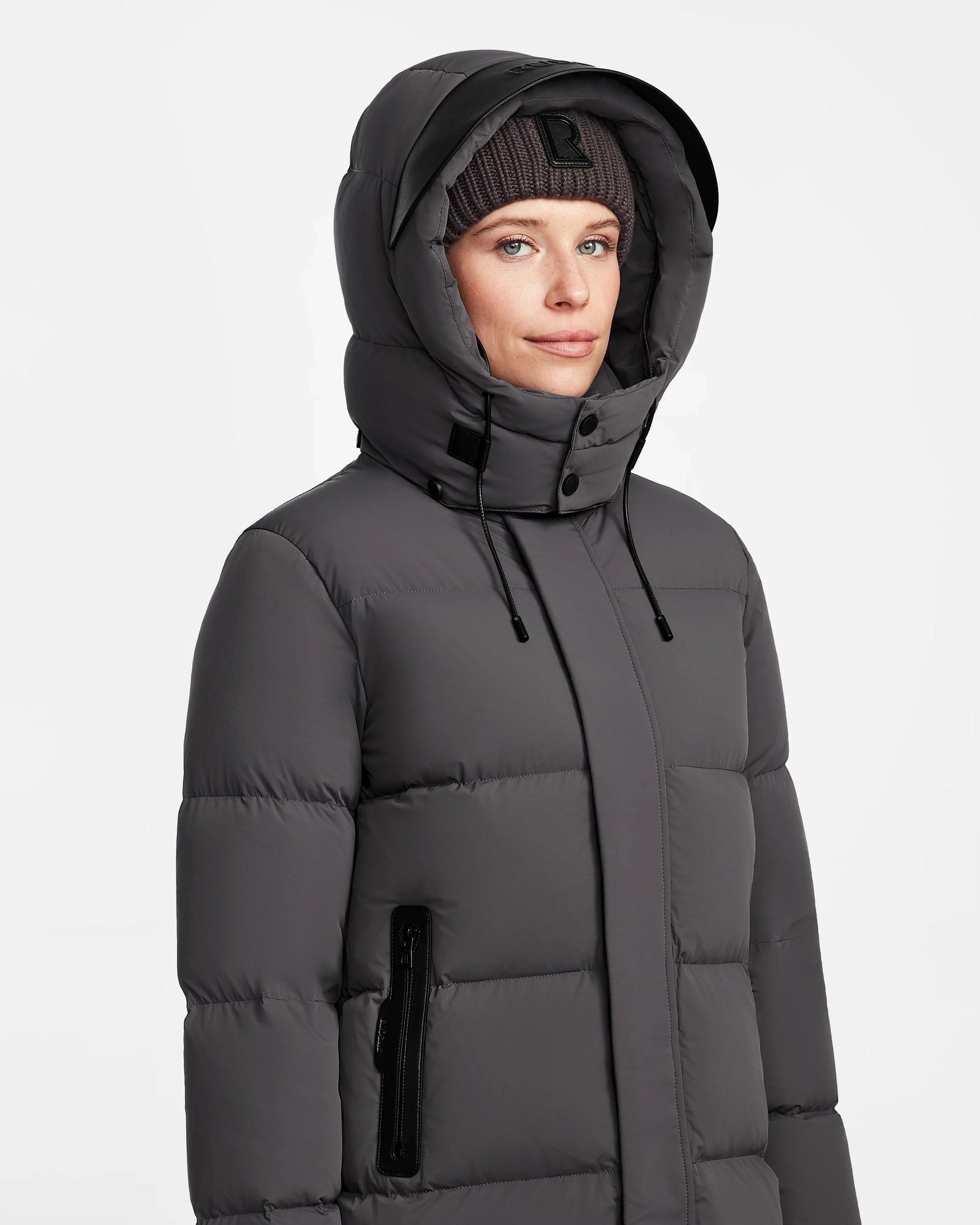 A woman wears the Rudsak JOELLE - 8124729 Grey quilted down coat with the hood up and a black knit beanie, gazing slightly to the side against a plain white background.