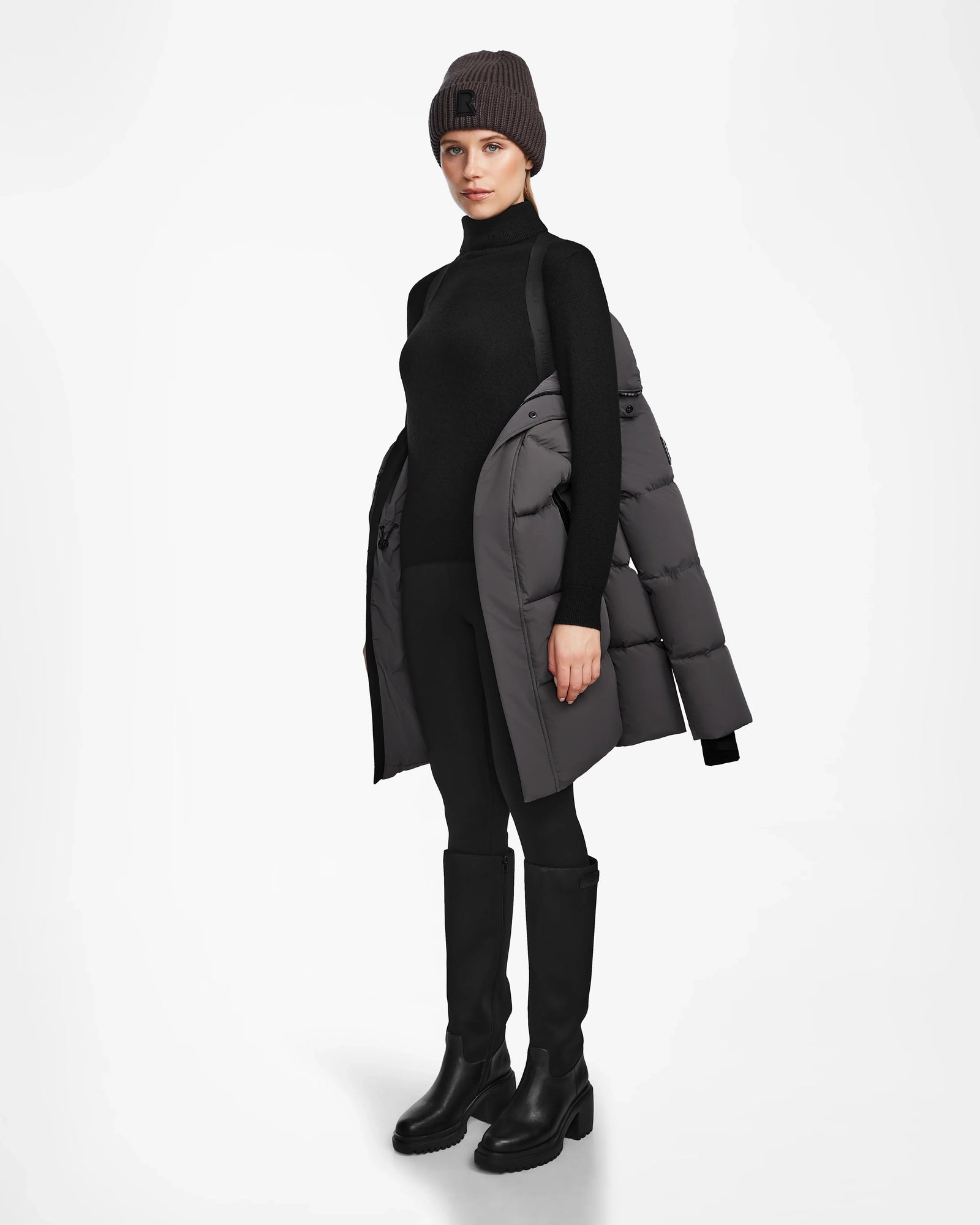 A woman in a black turtleneck, black pants, knee-high boots, a dark gray beanie, and the Rudsak JOELLE - 8124729 Grey RDS-certified down winter coat draped over her shoulders stands against a white background.