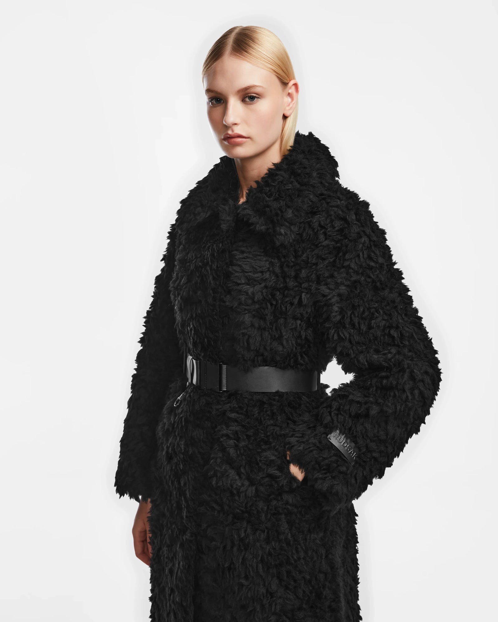 A woman with straight blonde hair models the Rudsak KAMILAH - 8124827 Black womens faux fur trench featuring a wide collar and vegan leather belt, posed against a white background.
