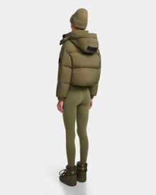 A person stands facing away, wearing the Rudsak KARLEE - 8124721 cropped down puffer in Loden Green with a matching beanie, fitted leggings, and boots. The quilted jacket pops against a plain white background.