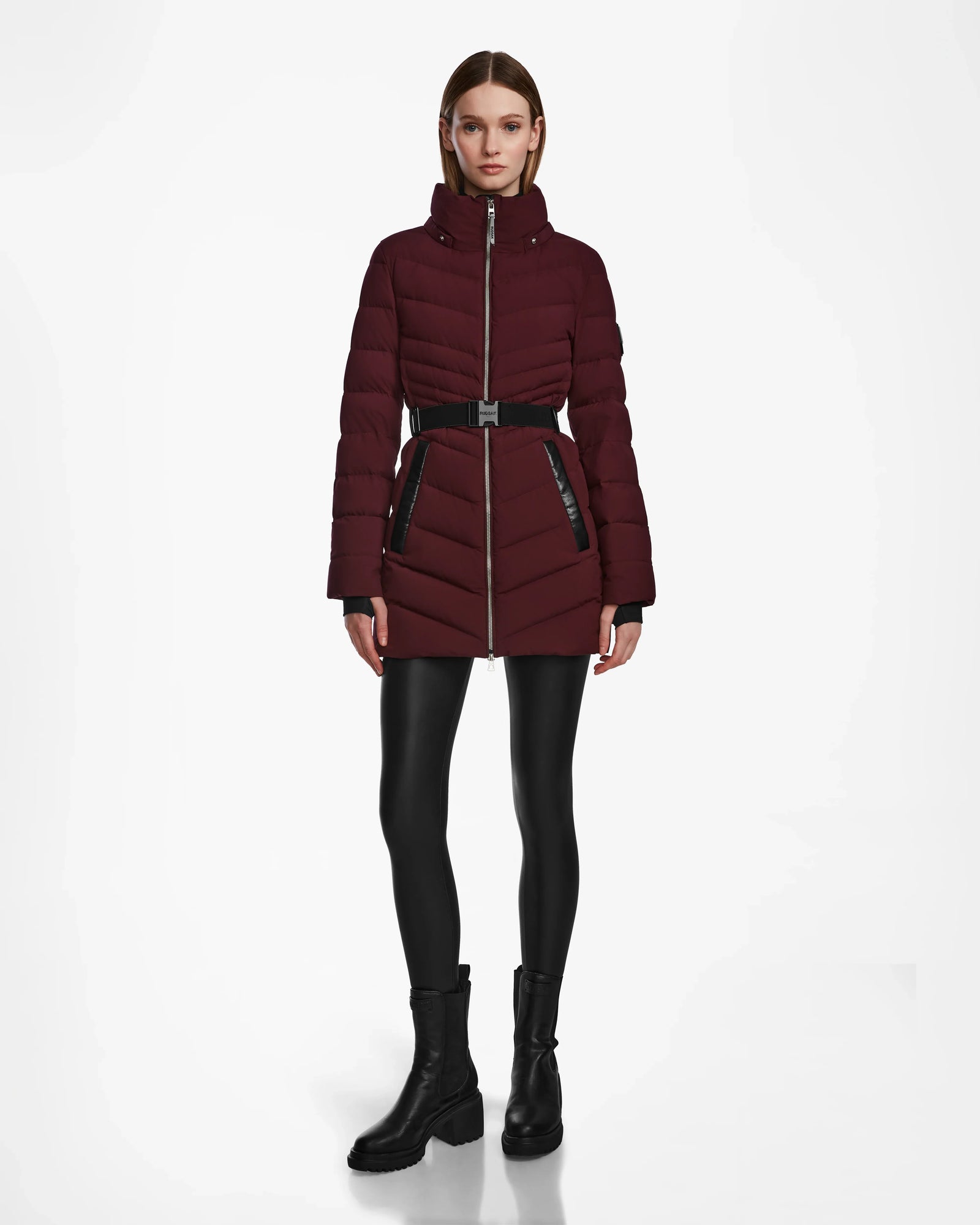 A woman faces forward against a white background, wearing the Rudsak MALAYA M - L123554 Wine belted womens down puffer jacket with herringbone quilting, black leggings, and black ankle boots.