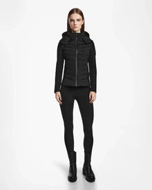 A woman stands facing forward in a Rudsak MALYA M - L123547 Black down quilted jacket, black leggings, and black ankle boots against a plain white background.