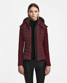 A woman with straight, light brown hair models the Rudsak MALYA M - L123547 Wine down quilted jacket over a black top and pants, standing against a plain white background.