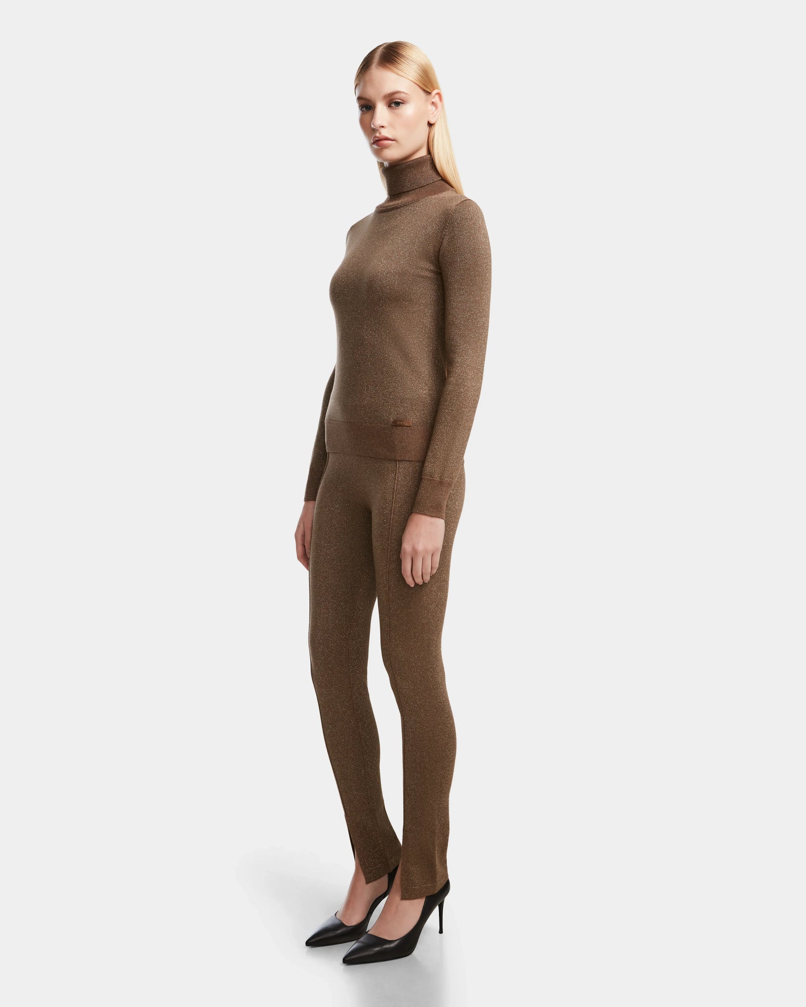 A woman wears the Rudsak MANSUR - 8524624 Antique Bronze textured long-sleeve turtleneck sweater and matching leggings with black heels, posing against a plain white background.