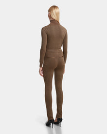 A woman with straight blonde hair faces away, wearing the Rudsak MANSUR - 8524624 Antique Bronze fitted turtleneck, chic ankle slit leggings, and black high heels against a plain white background.