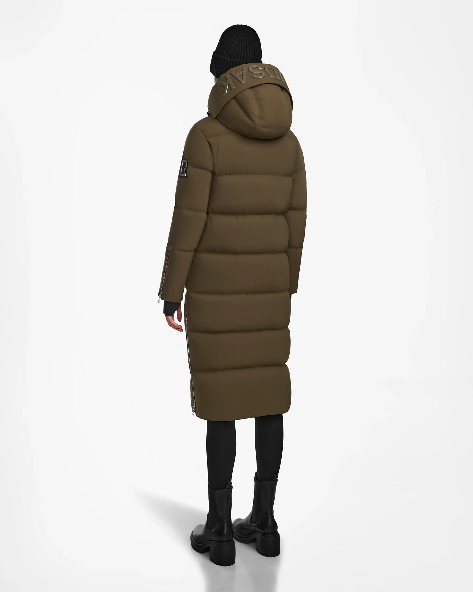 A person in a long Rudsak MAO M - 8123598 Army womens down puffer coat with hood, black beanie, leggings, and boots stands facing away against a white background.