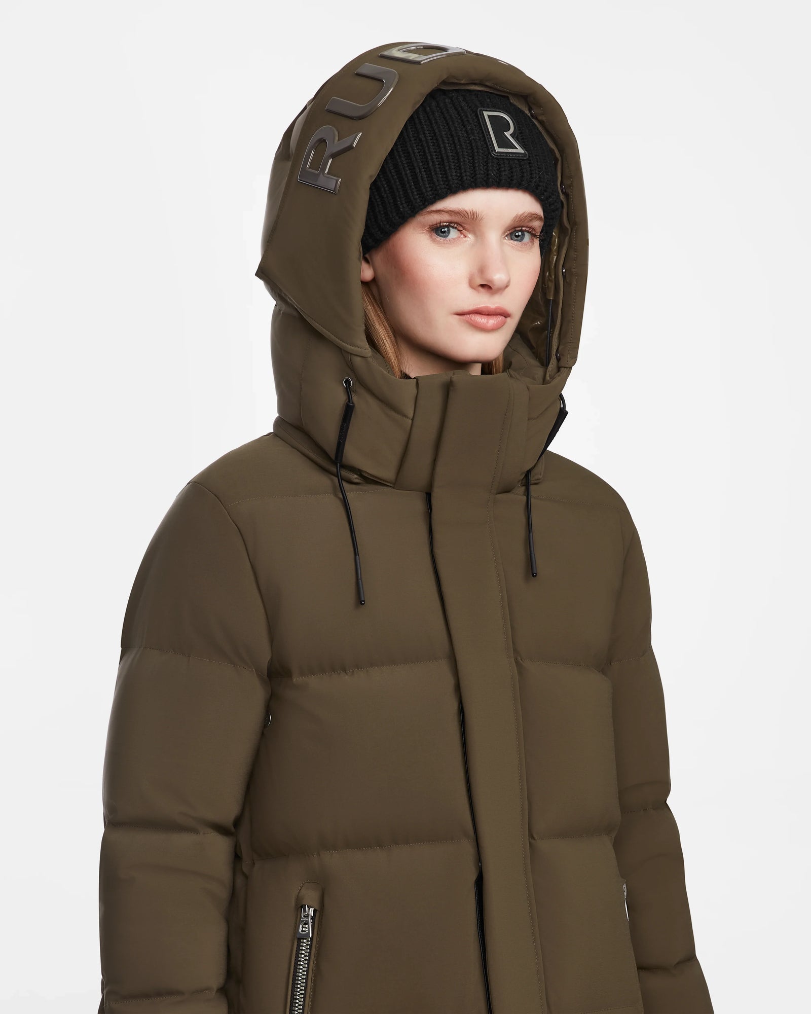 A woman stands against a plain white background wearing the Rudsak MAO M - 8123598 Army olive green winter coat with a hood and a black beanie, both featuring metallic RUB lettering.