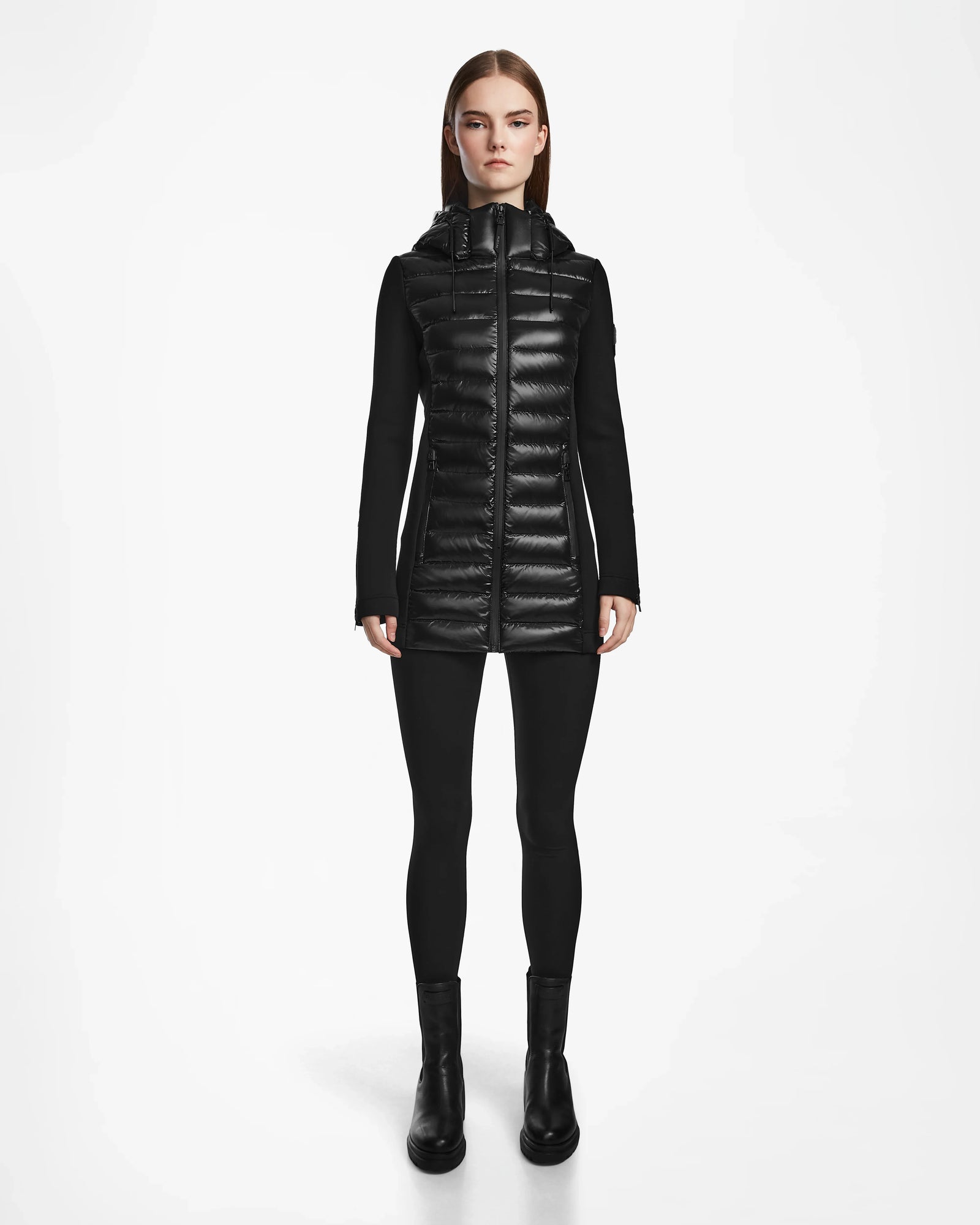 A woman stands facing forward against a plain white background, wearing the Rudsak MARRY - L121003 BLACK recycled down women’s puffer jacket with a hood, paired with black leggings and black ankle boots.