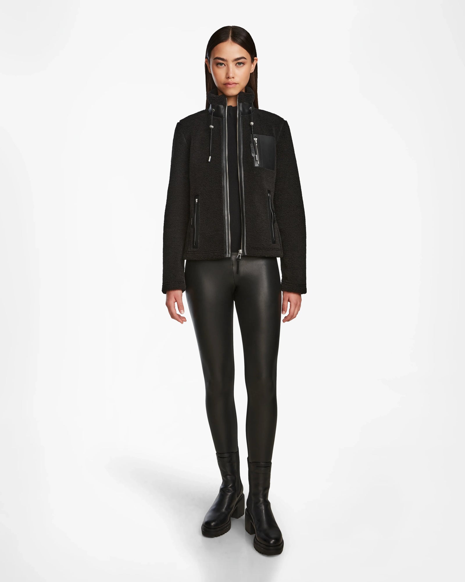 A woman faces forward wearing the Rudsak MILANA - 8524604 Black jacket, featuring vegan leather details, paired with black faux leather leggings and ankle boots against a plain white background.