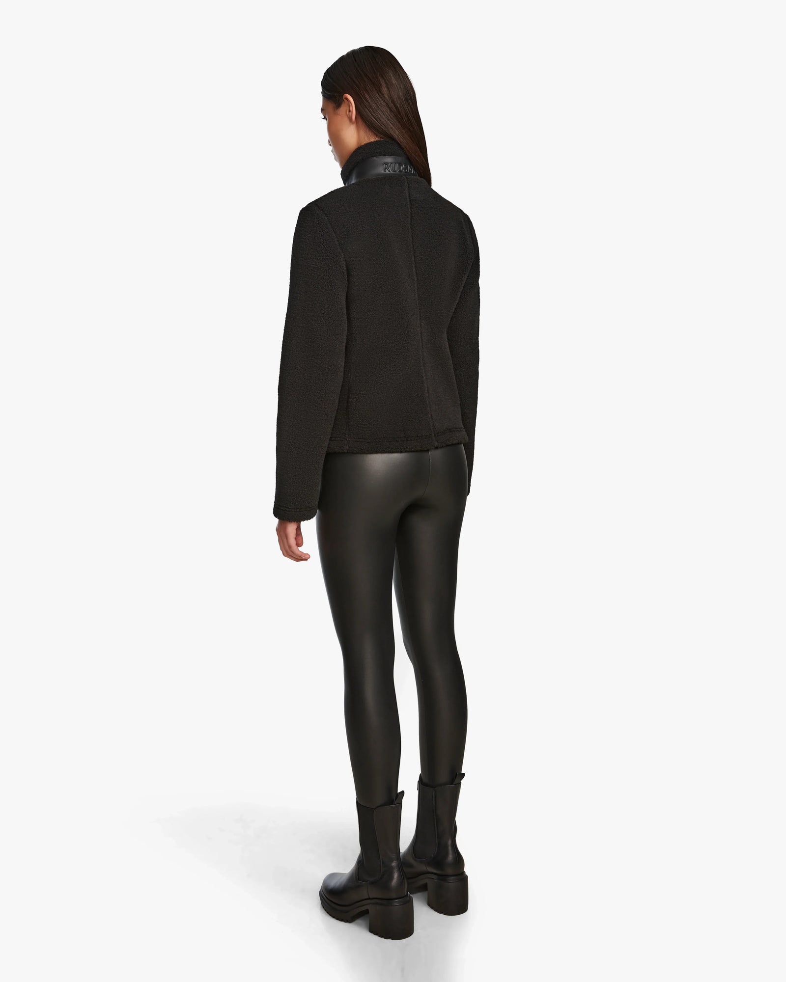 A person faces away, wearing the Rudsak MILANA - 8524604 Black womens teddy sweater with vegan leather details, paired with shiny leggings and black ankle boots, against a plain white background.