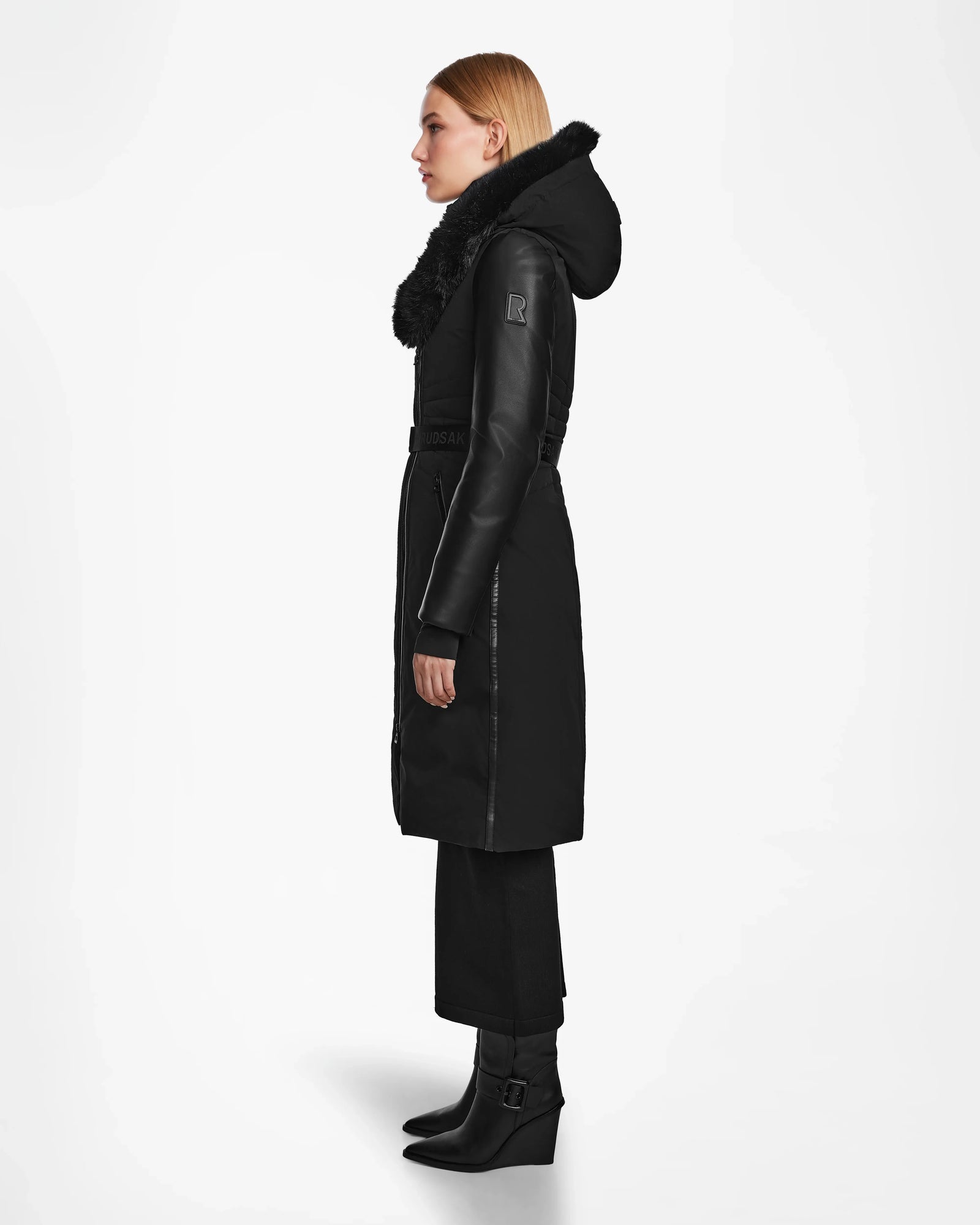 A woman stands in profile against a plain white background wearing the Rudsak MISALA - L124512 Black womens long down coat with a fur-trimmed hood, paired with black pants and black heeled boots.