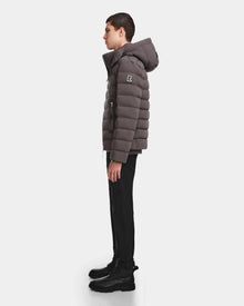 A person stands in profile wearing the Rudsak NAVAN - 6124551 Grey men’s quilted down puffer with a hood and “R” patch on the sleeve, paired with black pants and boots, against a plain white background.
