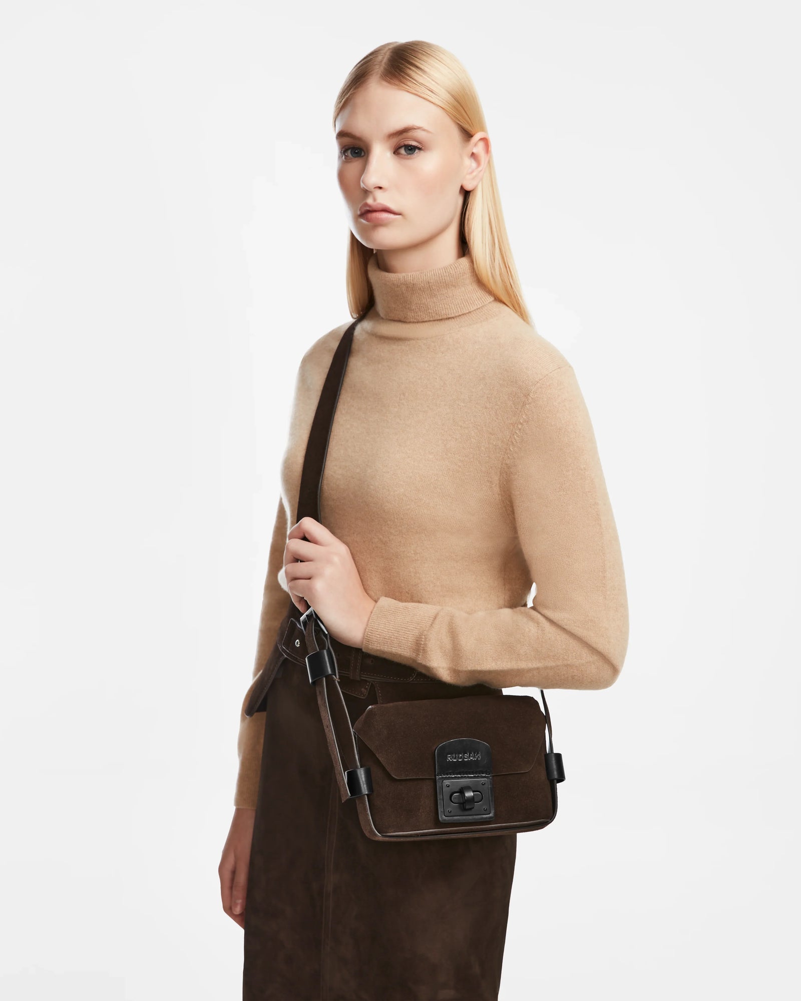 A woman with straight blonde hair in a beige turtleneck sweater and brown skirt stands against a white background, holding the Rudsak OFILIA S - 8324521 Espresso shoulder bag with a visible buckle and logo.