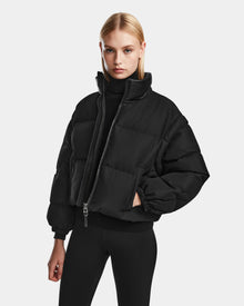 A woman with straight blonde hair models the Rudsak OPRAH - 8124708 Black womens cropped down puffer jacket with removable sleeves over a black turtleneck and leggings, standing against a plain light gray background.