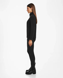 A woman with straight brown hair stands in profile, wearing the Rudsak ORCHID - 8524616 Black zip-up jacket with vegan leather trims, black leggings, and black boots against a plain white background.