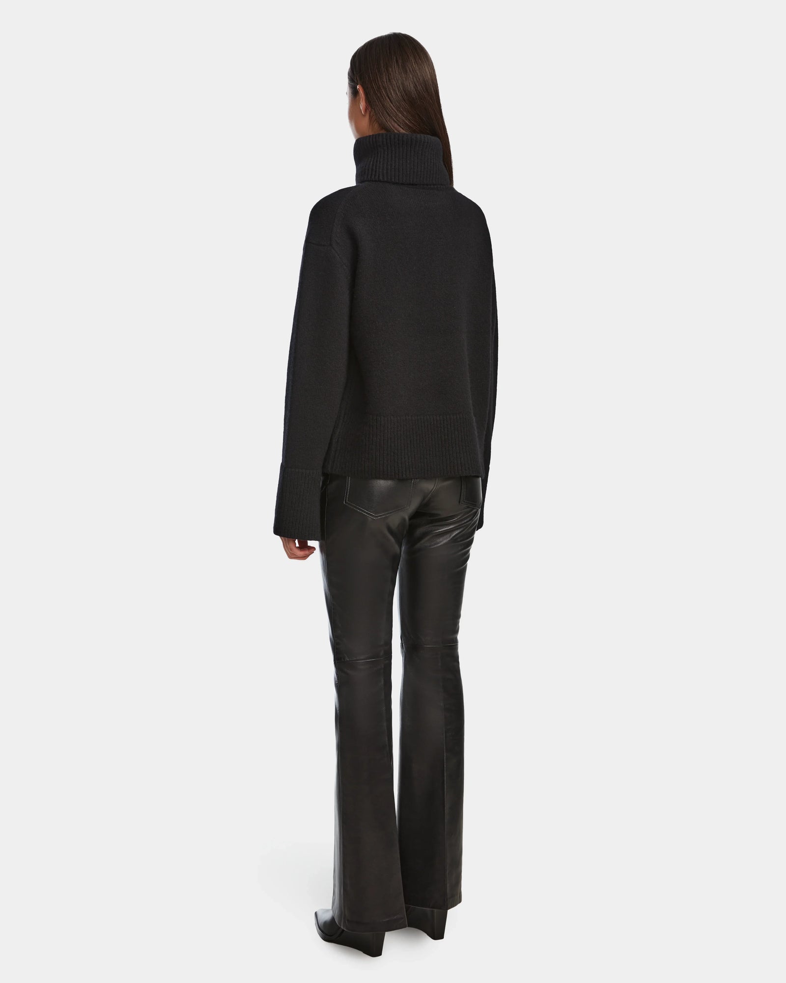 A person faces away, wearing Rudsaks PARMI - 8524608 Black womens knit turtleneck sweater and flared black leather pants, set against a plain white background.