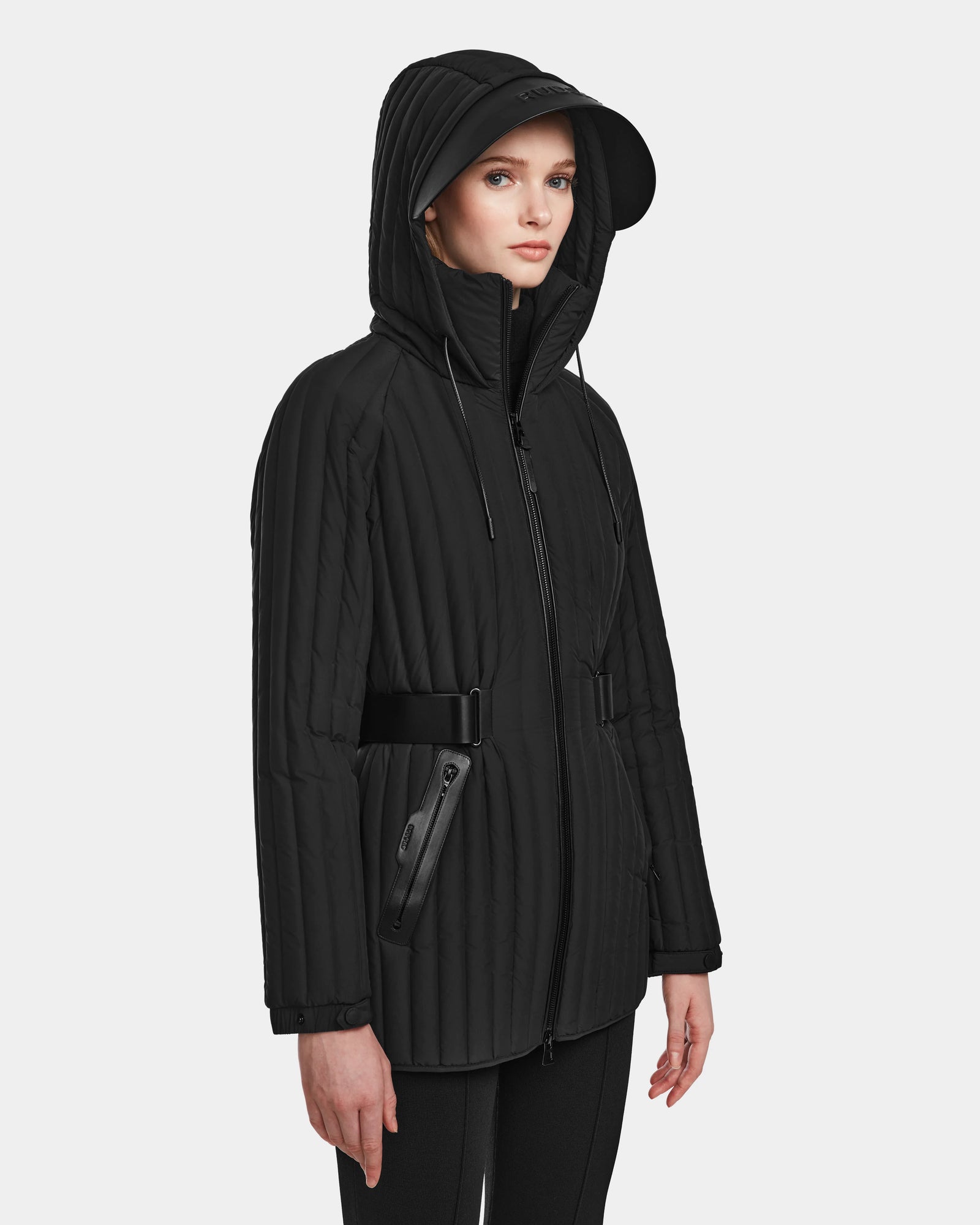 A woman models the Rudsak PETRAS - 8124725 Black quilted jacket with a hood, belted waist, and zippered pockets against a plain light background. She has a neutral expression and gazes slightly to the side.