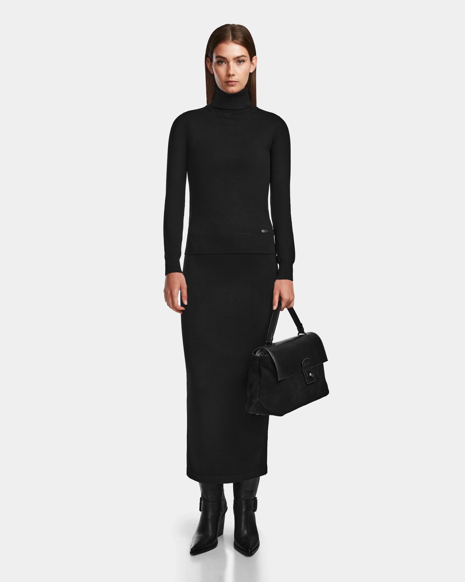 A woman faces forward against a white background, wearing the Rudsak RALDI - 8524605 Black womens knit turtleneck sweater with a matching long skirt and black boots, holding a black handbag by her side.