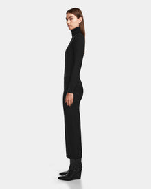 A woman stands in profile against a plain light background, wearing the RALDI - 8524605 Black knit turtleneck dress and black ankle boots with buckles by Rudsak.