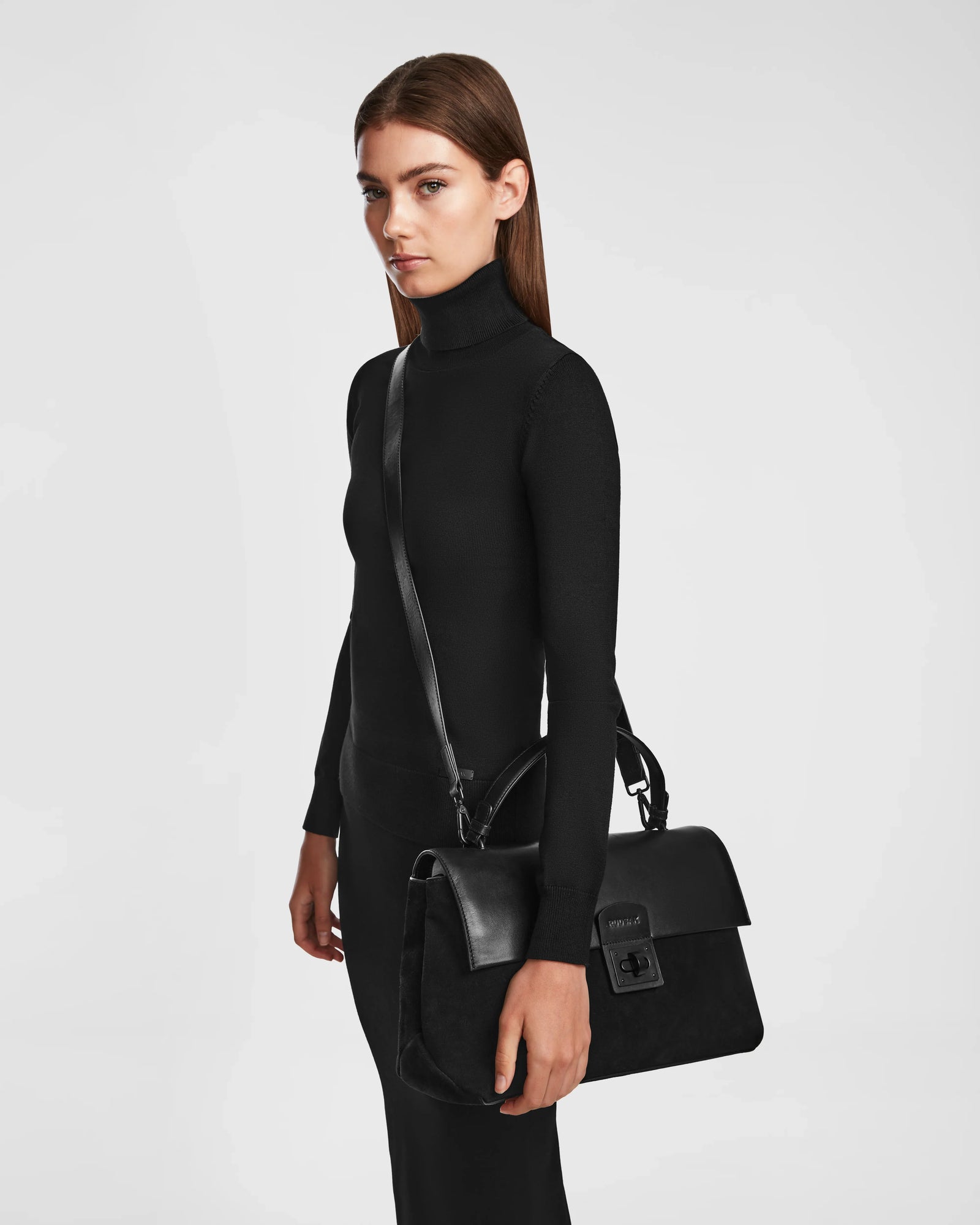 Against a plain white background, a woman with straight brown hair wears a fitted black turtleneck dress and holds the Rudsak ESMA - 8324519 Black suede and leather handbag featuring a top handle and removable shoulder strap.