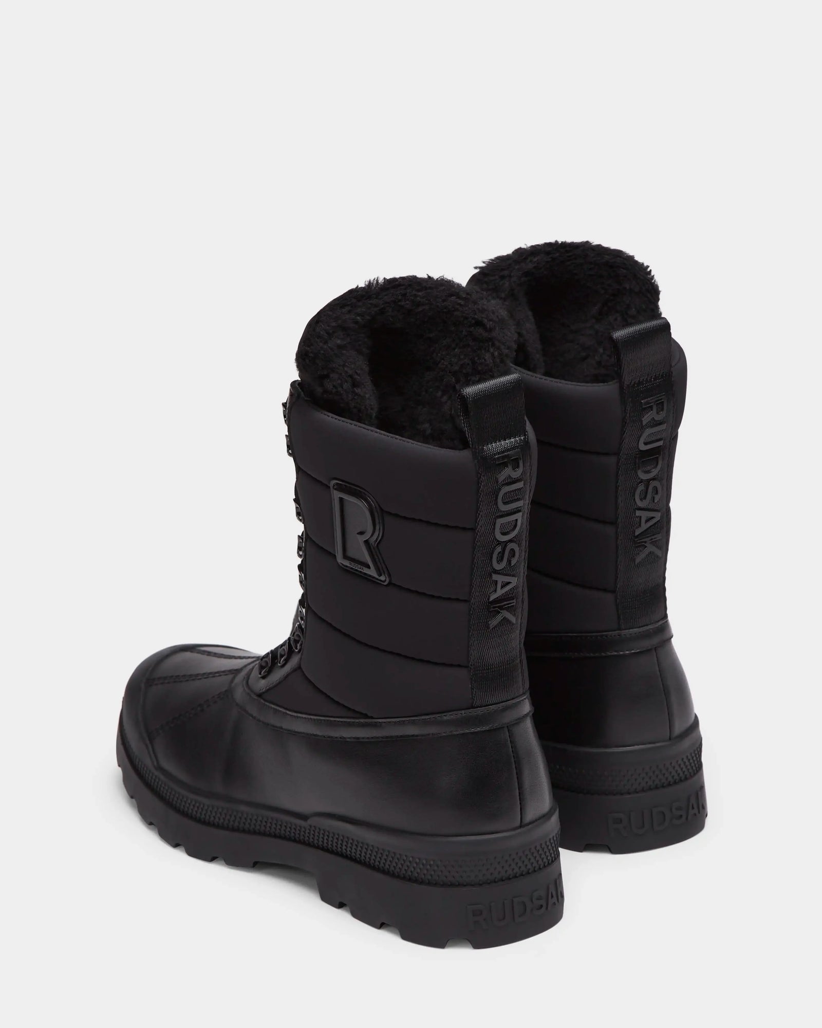 The Rudsak REAGAN - 6224501 Black men’s winter boots feature thick soles, lace-up fronts, soft furry linings, a back pull tab, and the Rudsak name embossed vertically for a stylish finish.