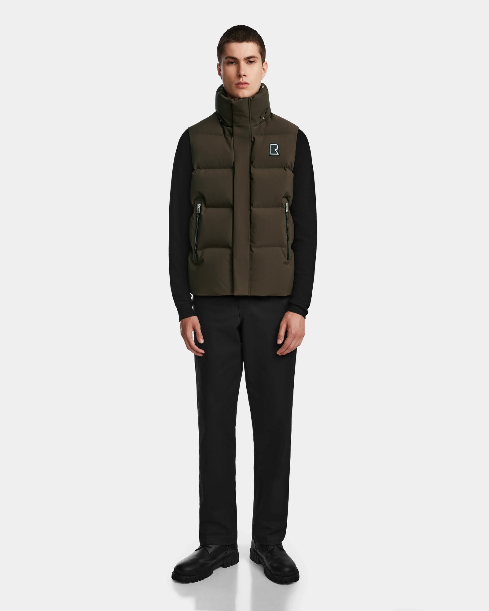 A young man faces forward, wearing the Rudsak REN M - 9123512 Army M weather-resistant olive green puffer vest over a black long-sleeve shirt, black pants, and black shoes, set against a plain light gray background.