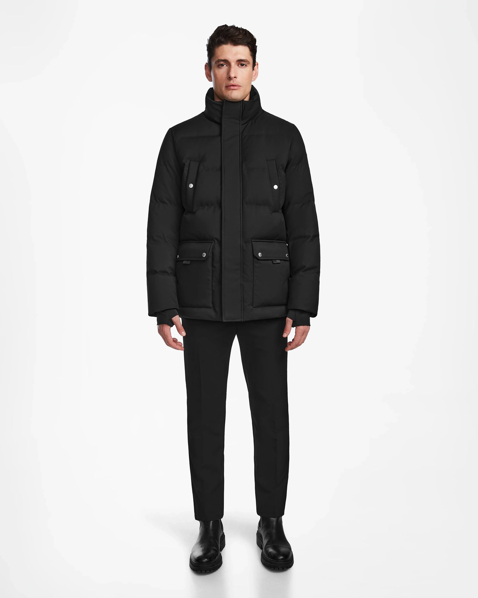A man stands facing forward wearing the Rudsak RICKY - M123502 Black men's quilted down parka with large pockets, black pants, and black boots against a plain white background.