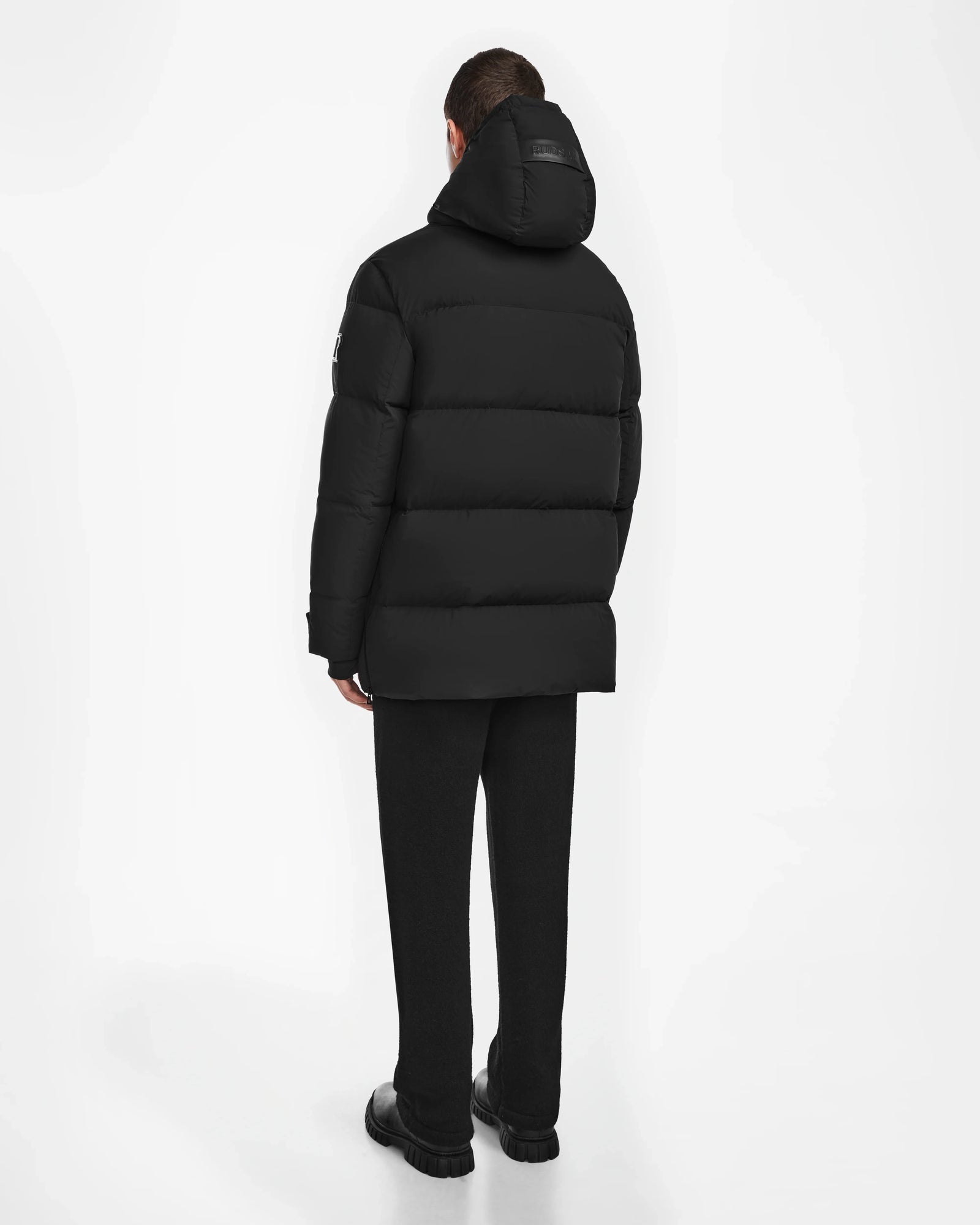 A person faces away from the camera, wearing the Rudsak ROKO - 6124510 Black mens quilted down parka with RDS-certified insulation, paired with black pants and shoes, set against a plain white background.