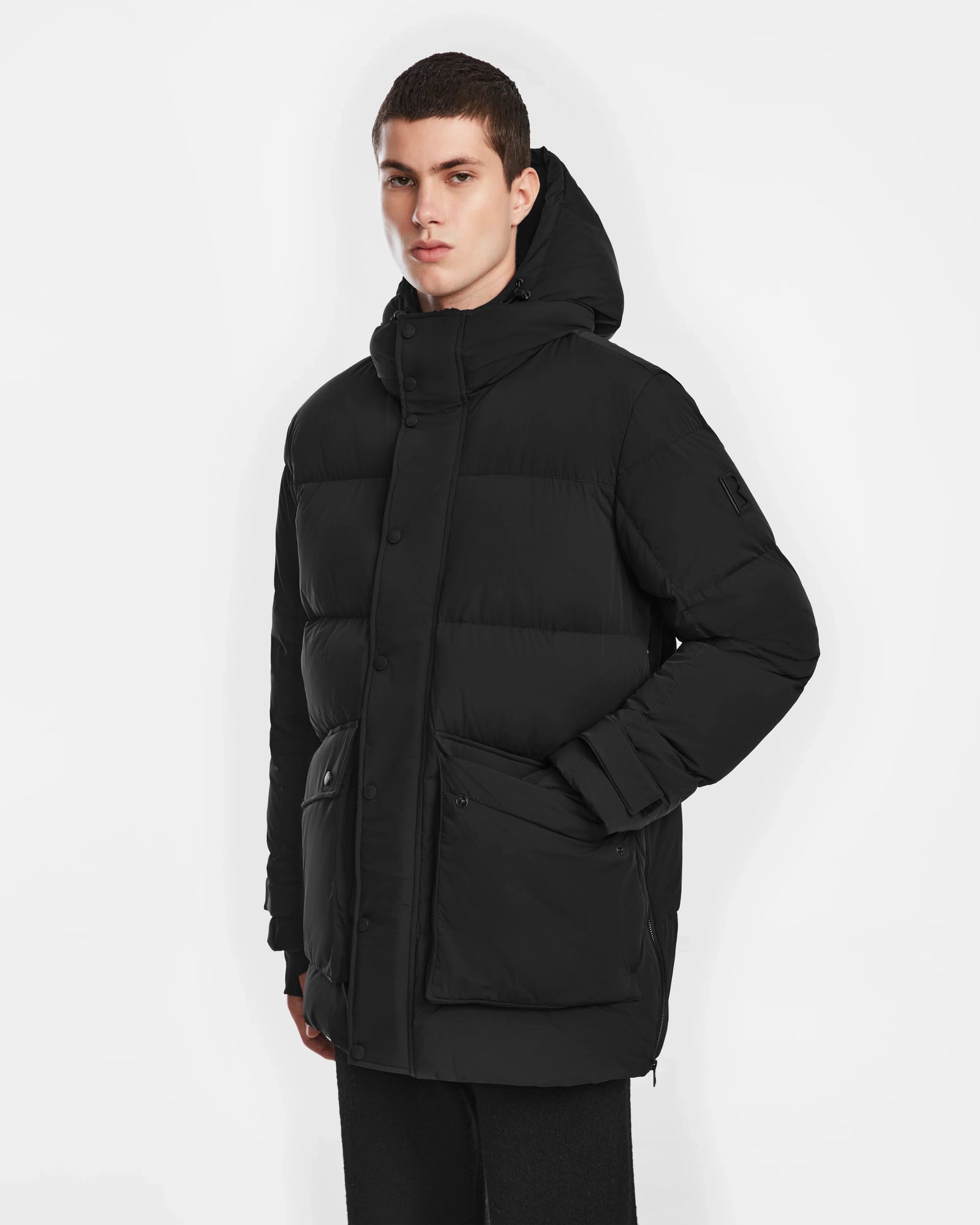 A person with short dark hair wears the Rudsak ROKO - 6124510 Black mens quilted down parka with RDS-certified insulation. Buttoned up with large pockets, they stand against a white background and look forward neutrally.