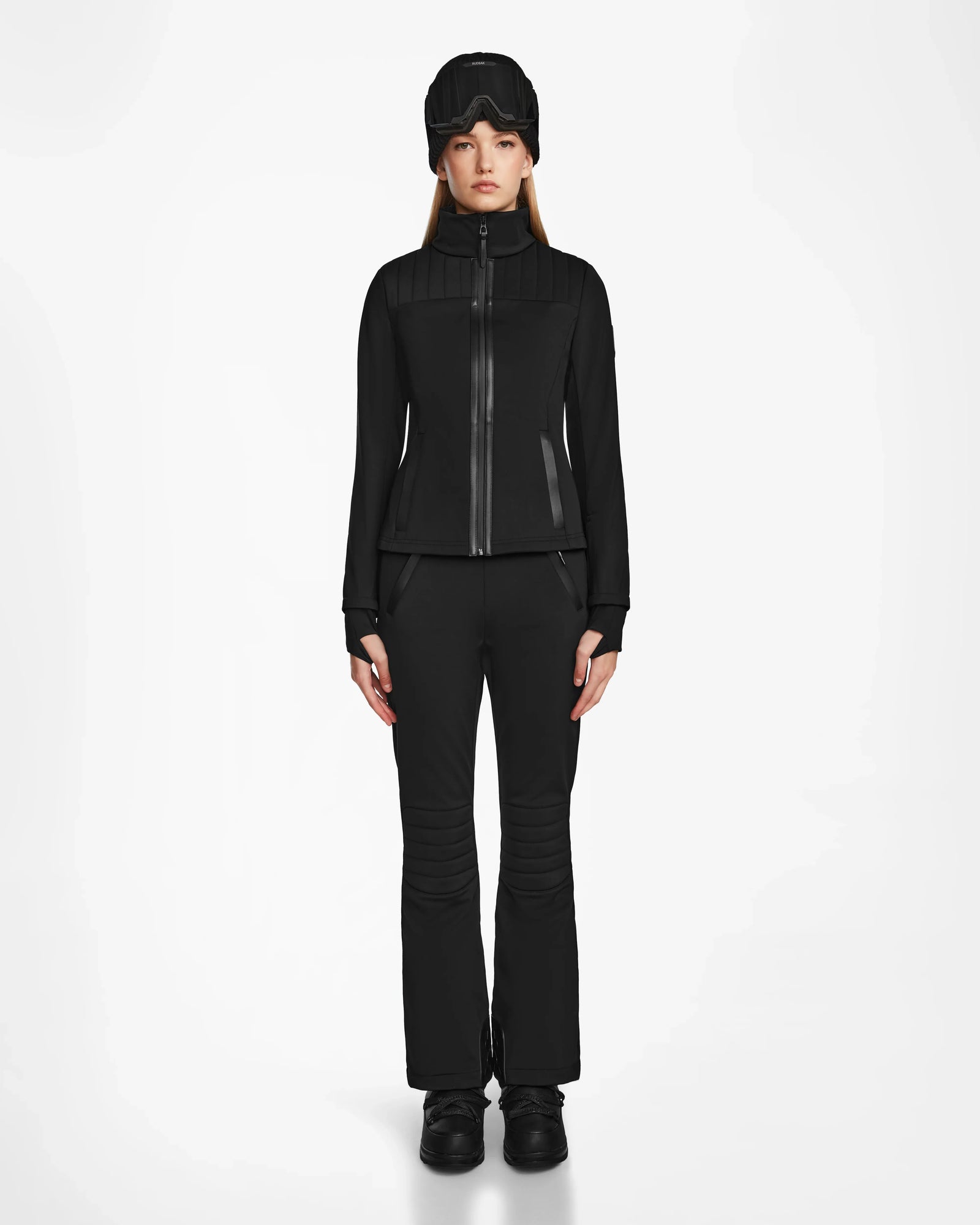 A woman faces forward wearing the Rudsak ROSALIE - 8124784 Black fitted ski jacket with matching black pants, boots, and a black winter hat, set against a plain white background.