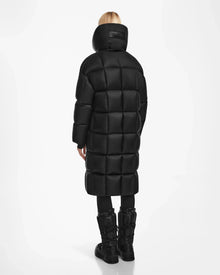 A person with blonde hair is seen from behind wearing the Rudsak SASH - 8124536 Black womens long down puffer coat, paired with black pants and boots, against a plain white background.