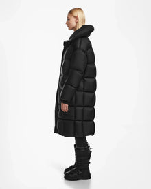 A person with straight blonde hair, wearing the Rudsak SASH - 8124536 Black womens long down puffer coat, stands in profile against a plain white background.
