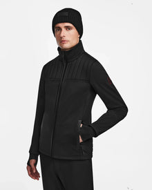 A man wears the Rudsak TREVOR - 6124519 Black mens ski jacket, black pants, and a knit beanie, standing with one hand in his pocket against a white background to highlight its technical performance and style.