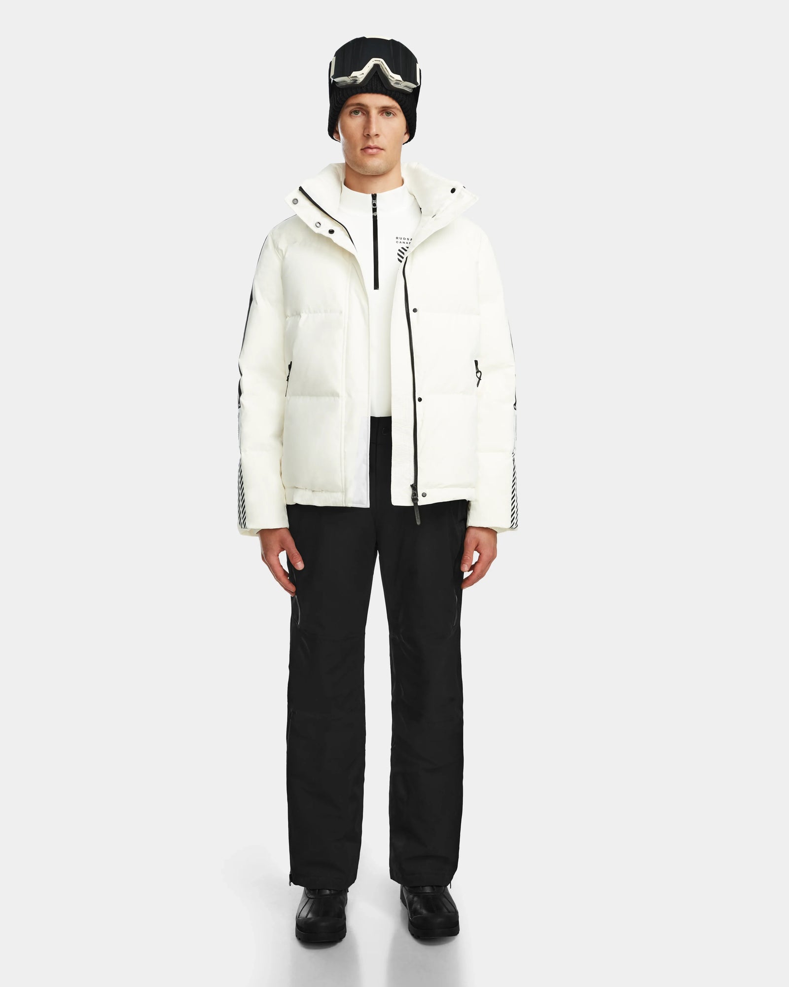 A person stands facing forward against a plain light background, wearing the Rudsak USHER - 6124516 Optic White men’s down ski jacket with RDS-certified insulation, black pants and beanie, white zip-up top, black shoes, and ski goggles.
