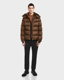 A person faces forward wearing the Rudsak VINCE S - 6124560 Antique Bronze men’s quilted down puffer over a tan sweater, black pants, and black shoes, set against a plain white background.