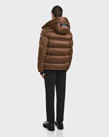A person stands facing away, wearing the Rudsak VINCE S - 6124560 Antique Bronze mens quilted down puffer jacket with a hood, along with black pants and shoes, set against a plain white background.