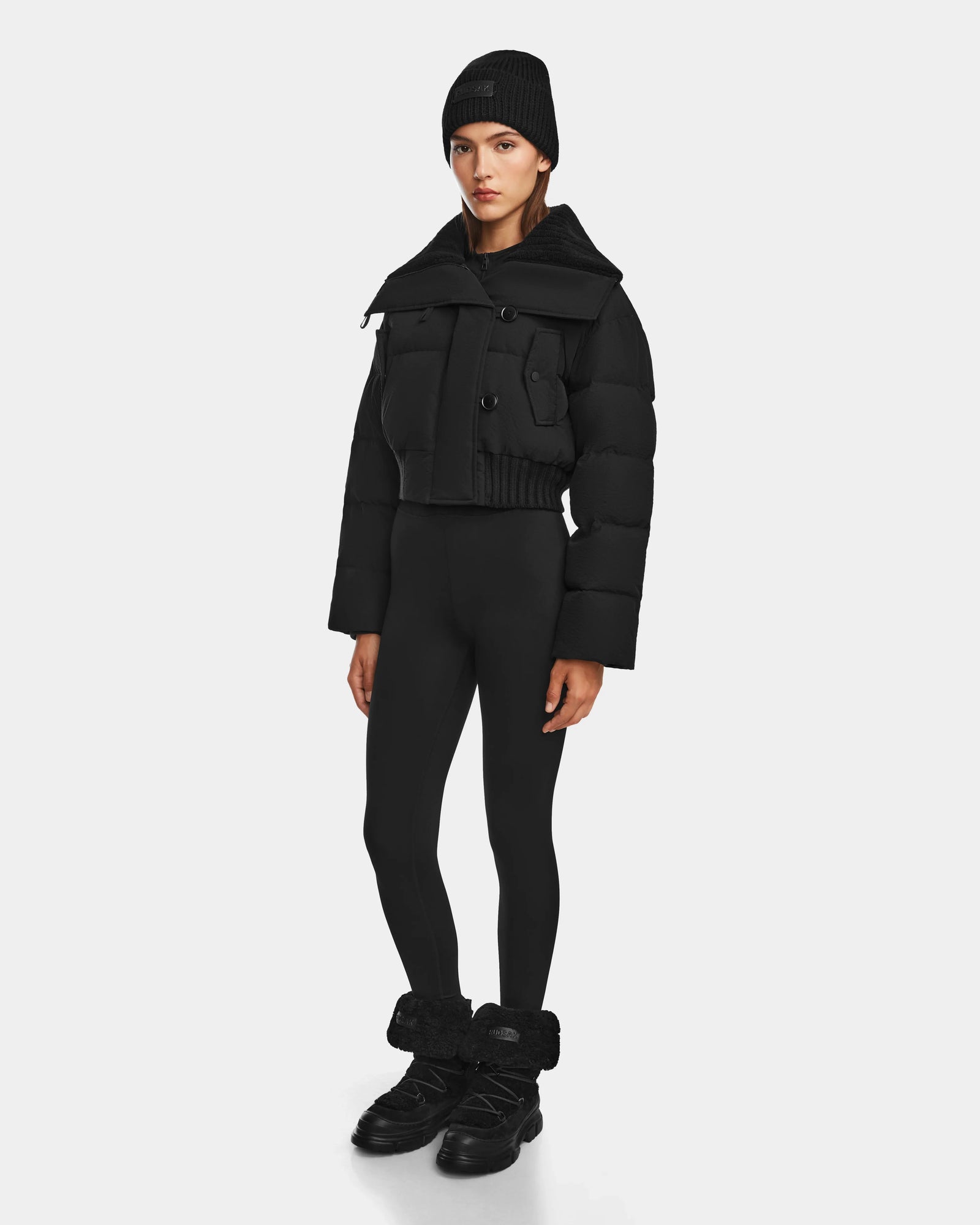 A person stands facing forward, wearing Rudsaks YVETTE - 8124710 Black crop down puffer jacket, paired with a black knit beanie, black leggings, and black shoes with fuzzy ankle cuffs, highlighting chic winter style against a plain white background.