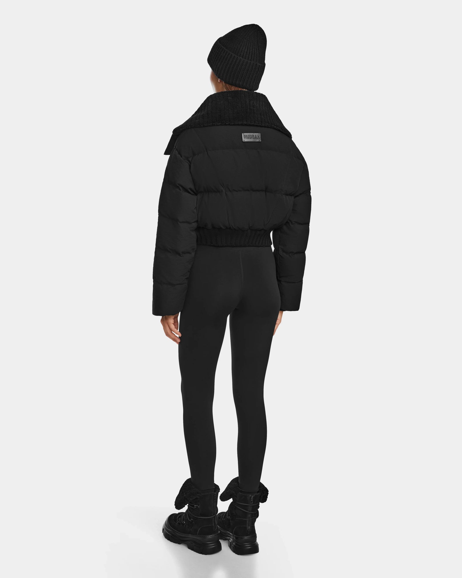 A person models the Rudsak YVETTE - 8124710 Black women’s crop down puffer, paired with black leggings and bow boots, standing against a plain white background.