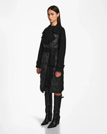 A woman with straight brown hair and a neutral expression models the Rudsak ZOE - 8124711 Black womens long quilted down trench coat with a wide collar and belted waist, paired with knee-high black heeled boots, against a white background.