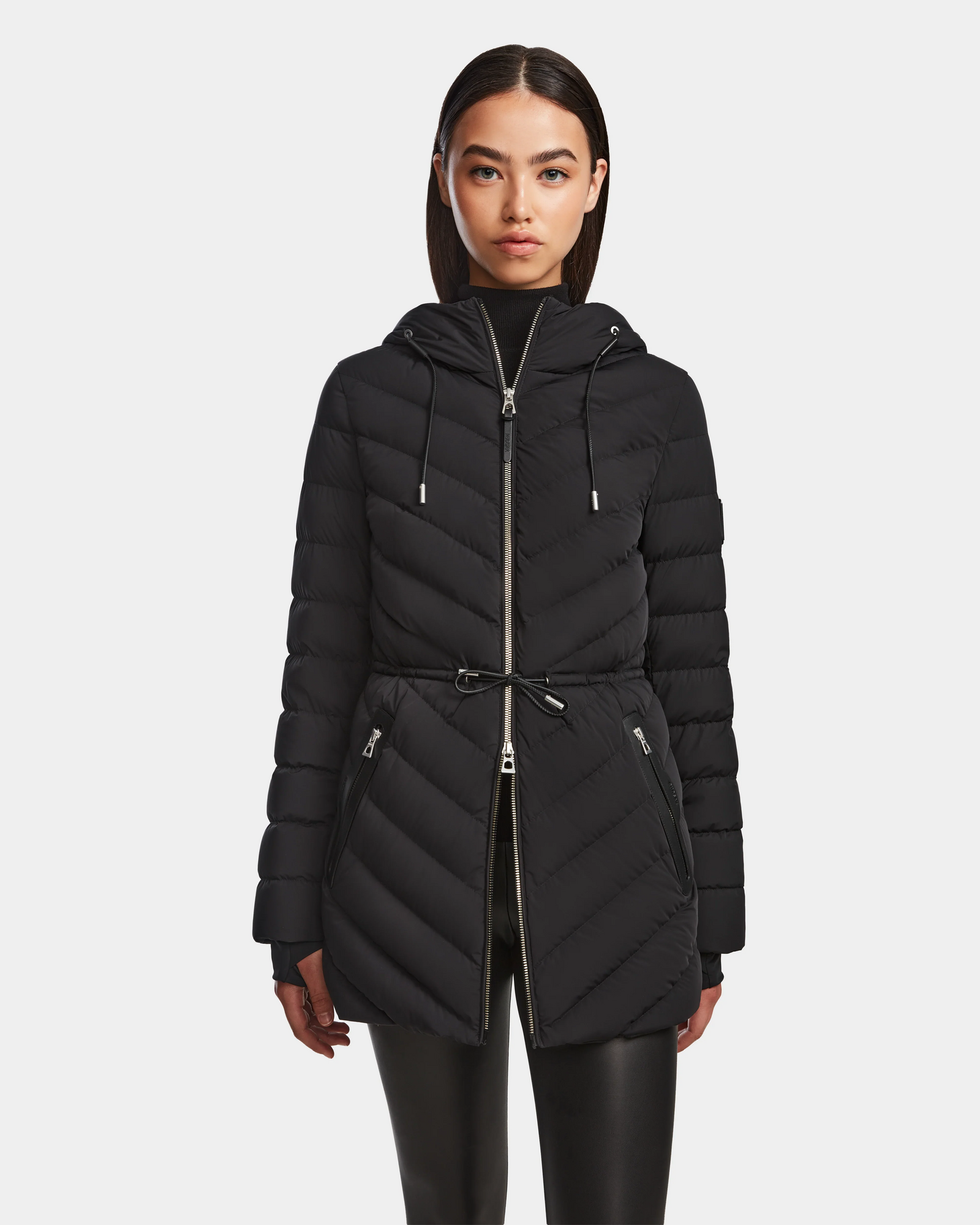 A woman with straight dark hair wears the Rudsak FRANCINE - 8124722 Black quilted puffer hooded jacket, featuring a front zipper, drawstring waist, and zippered pockets, paired with black pants against a plain white background.