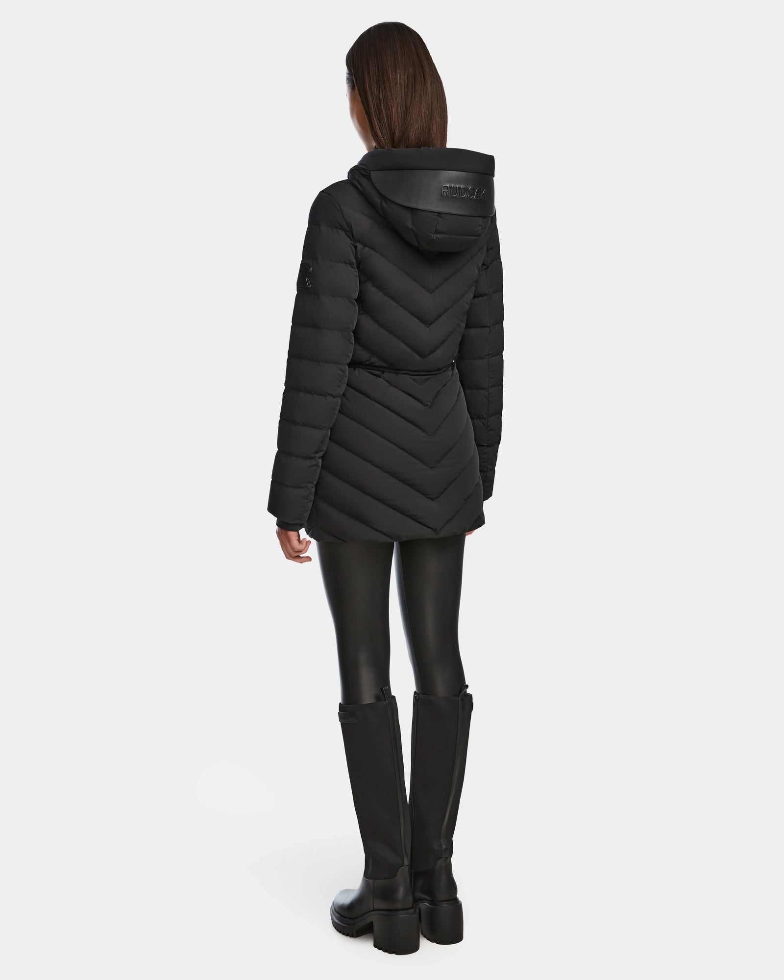 A person is seen from the back wearing the Rudsak FRANCINE - 8124722 Black quilted puffer jacket with a hood, paired with black leggings and tall black boots, set against a plain white background.