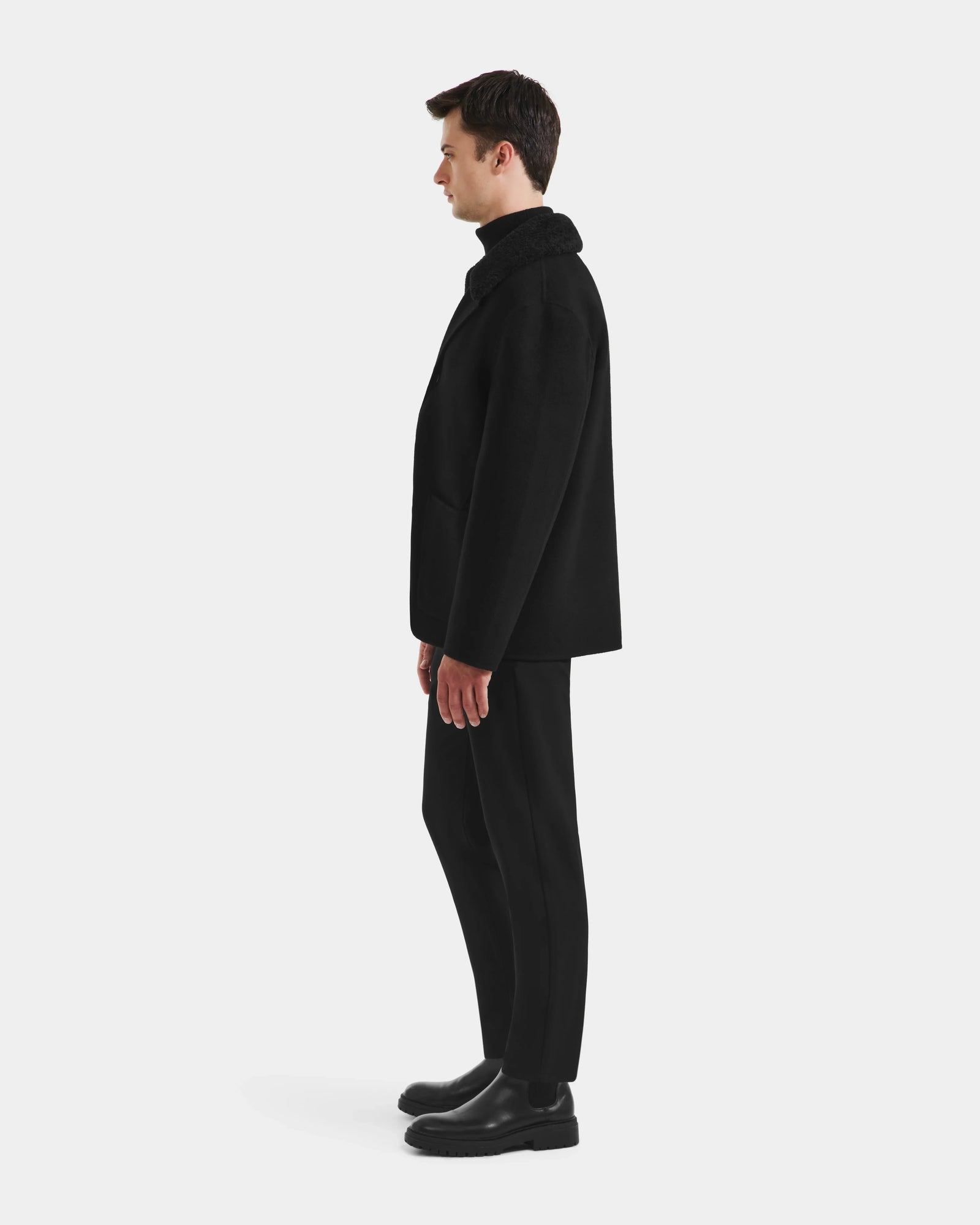 A man stands in profile facing left, wearing the Rudsak AIDEN - 6125558 Black men’s wool-blend coat with black pants and shoes, set against a plain white background.